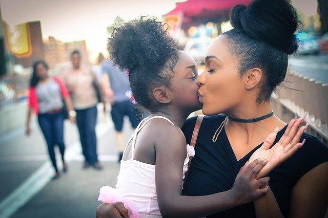 A woman holds and kisses her young daughter in the middle of a busy city sidewalk.