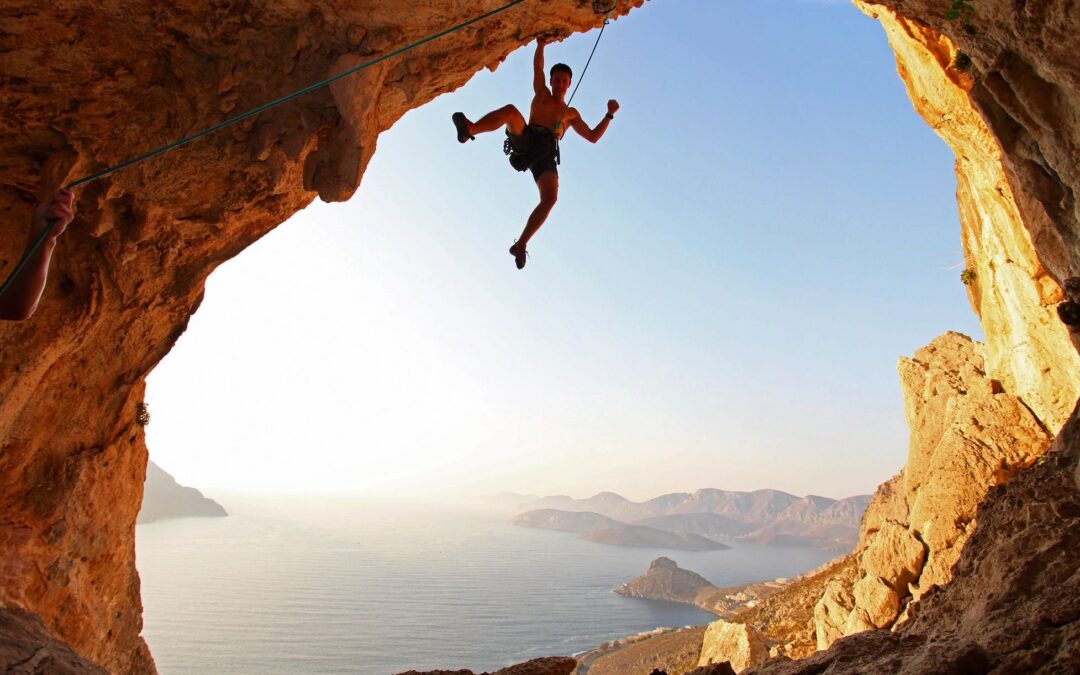 A man, rock climbing, dangles from a cliff with one hand.